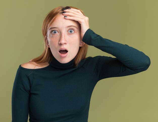 excited young redhead ginger girl with freckles puts hand on head isolated on olive green wall with copy space