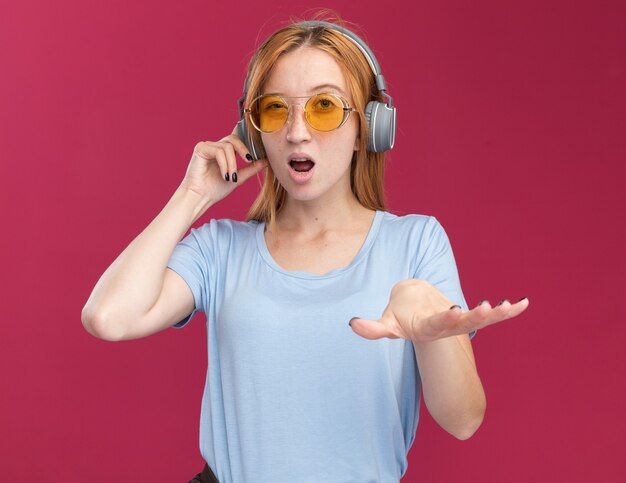 Excited young redhead ginger girl with freckles in sun glasses and on headphones holds hand open isolated on pink wall with copy space