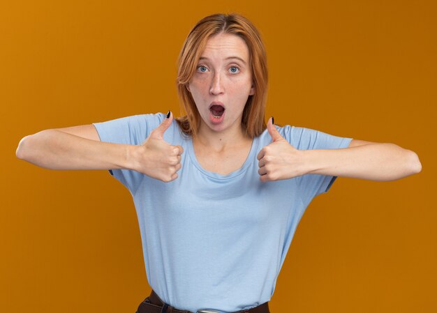 Excited young redhead ginger girl with freckles thumbing up of two hands isolated on orange wall with copy space