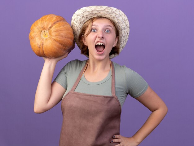 Free photo excited young slavic female gardener wearing gardening hat holding pumpkin