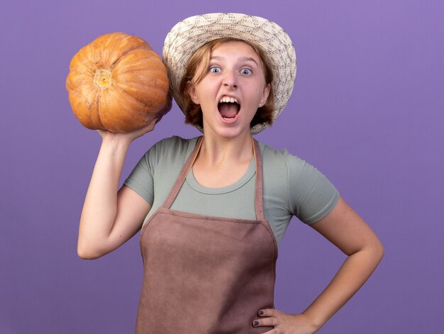 Excited young slavic female gardener wearing gardening hat holding pumpkin