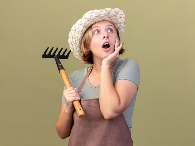 Excited young slavic female gardener wearing gardening hat puts hand on face and holds rake looking at side on olive green