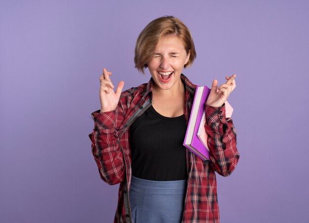 Excited young slavic student girl stands with closed eyes crossing fingers holding book and notebook 