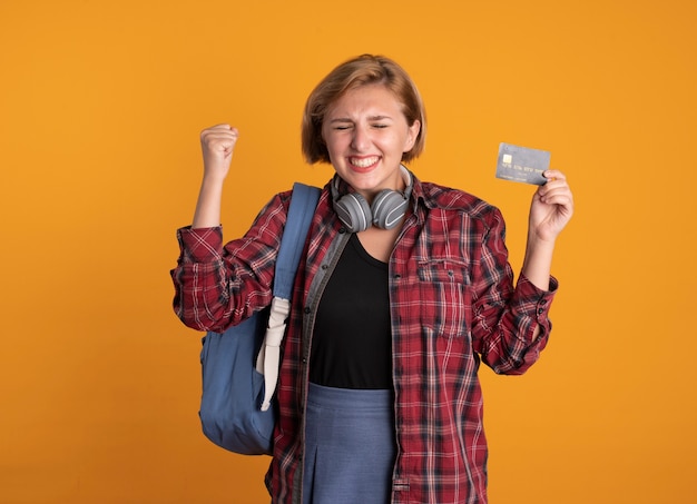 Возбужденная молодая славянская студентка с наушниками в рюкзаке стоит с закрытыми глазами, поднимая кулак, держа кредитную карту