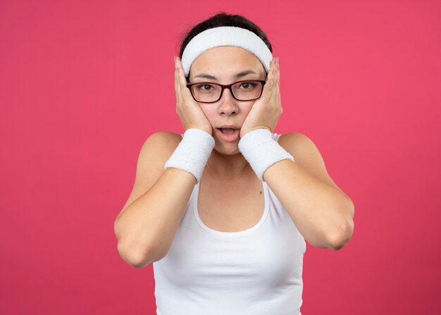 Возбужденная молодая спортивная девушка в оптических очках с повязкой на голову и браслетами кладет руки на лицо