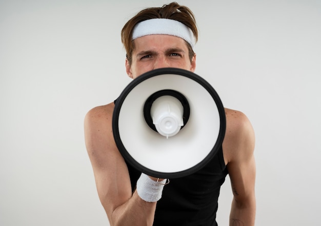 Excited young sporty guy wearing headband and wristband speaks on loudspeaker isolated on white