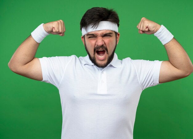 Excited young sporty man wearing headband and wristband showing strong gesture isolated on green