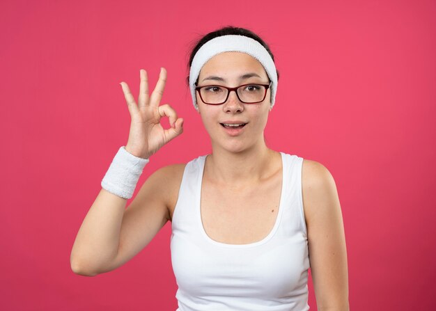 Возбужденная молодая спортивная женщина в оптических очках с повязкой на голову и браслетами жестами показывает знак рукой на розовой стене