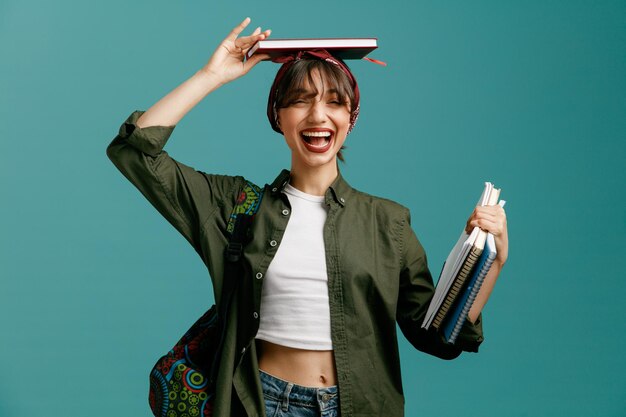 excited young student girl wearing bandana and backpack holding note pads looking at camera putting one of note pads on head isolated on blue background