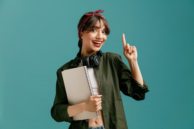 Excited young student girl wearing bandana and headphones around neck holding large note pad looking at camera pointing up isolated on blue background