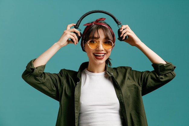 Free photo excited young student girl wearing bandana sunglasses holding headphones above head looking at camera isolated on blue background