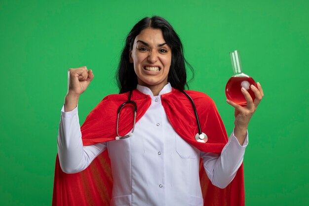 Возбужденная молодая девушка-супергерой в медицинском халате со стетоскопом держит стеклянную бутылку химии, наполненную красной жидкостью, показывая жест да, изолированный на зеленой стене