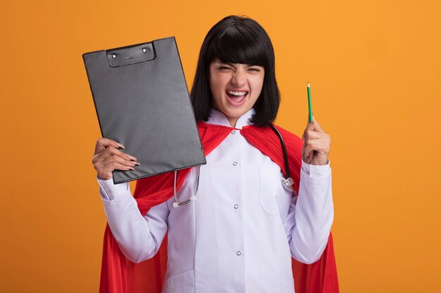 Excited young superhero girl wearing stethoscope with medical robe and cloak holding clipboard with pencil isolated on orange