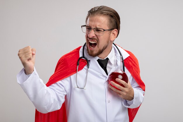 Excited young superhero guy wearing medical robe with stethoscope and glasses holding chemistry glass bottle filled with red liquid showing yes gesture isolated on white background