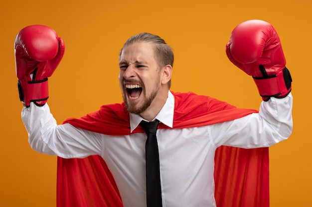 Excited young superhero guy wearing tie and boxing gloves raising hands isolated on orange background