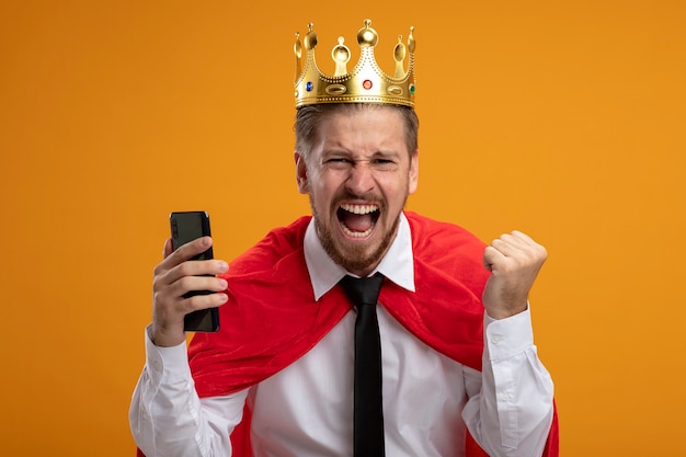 Free photo excited young superhero guy wearing tie and crown holding phone showing yes gesture isolated on orange background