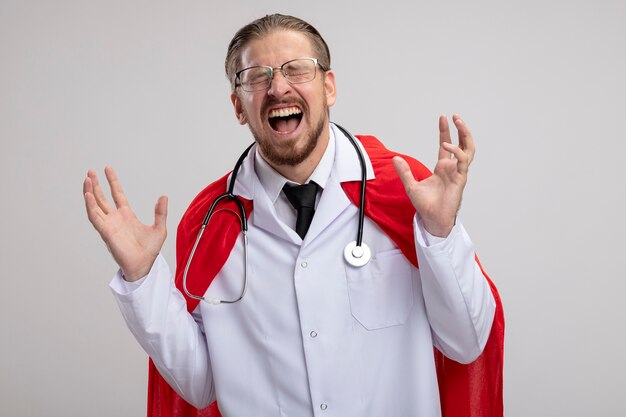 Excited young superhero guy with closed eyes wearing medical robe with stethoscope and glasses isolated on white background
