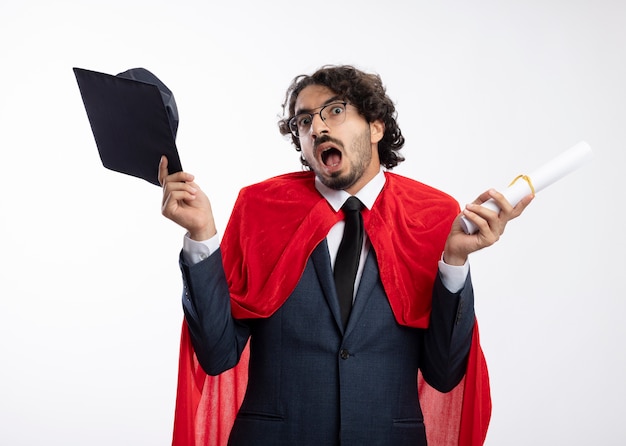 Возбужденный молодой супергерой в оптических очках в костюме с красным плащом держит выпускную кепку и диплом, изолированные на белой стене
