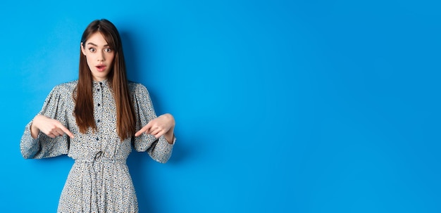 Excited young woman in dress pointing fingers down at logo looking intrigued at camera standing on b