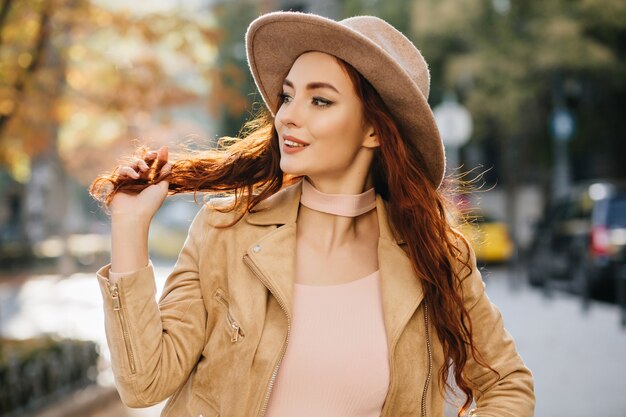 Excited young woman in elegant attire looking away and playing with ginger hair