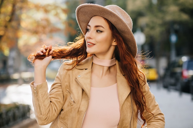 Excited young woman in elegant attire looking away and playing with ginger hair