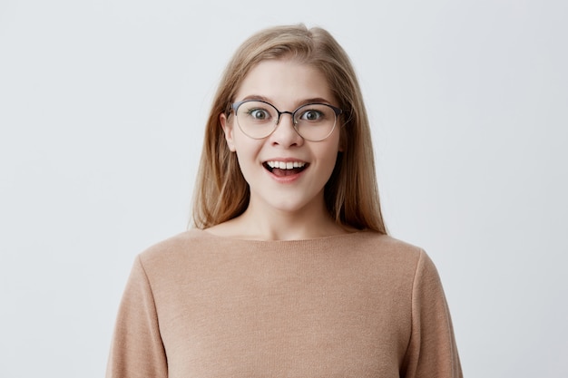 Excited young woman of European appearance wearing brown loose sweater, with blonde hair, in eyeglasses, smiling broadly in amazement, demonstrating her perfect white teeth. Youth and happiness