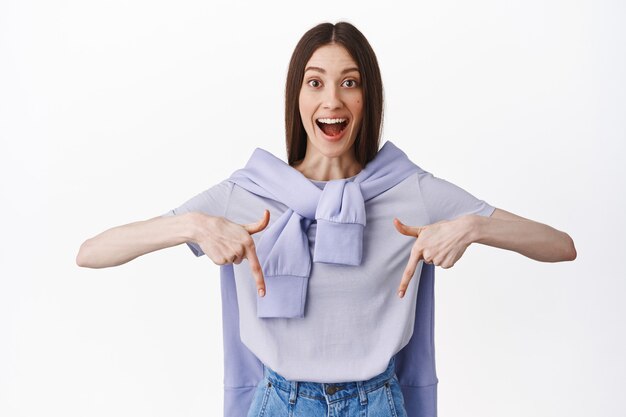 Excited young woman found good deal, pointing at promotional text with fascinated face, gasping and smiling happy, showing advertisement bellow, white wall