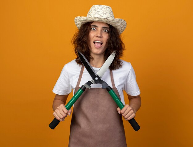 Excited young woman gardener in uniform wearing gardening hat holding clippers isolated on orange