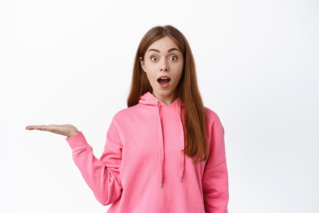 Excited young woman gasping amazed saying wow holding empty copy space on palm spread hand sideways display product standing over white background