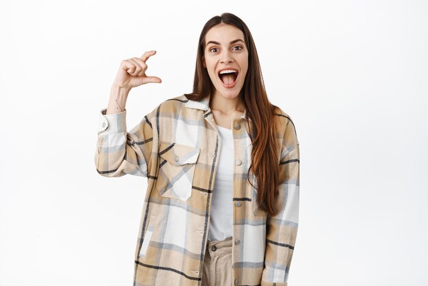 Excited young woman holding something little and awesome scream from joy and amazement as hold small size object on copyspace standing over white background