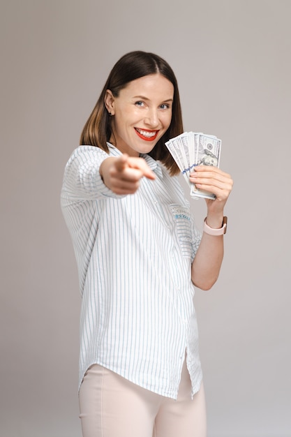 회색 벽 위에 고립된 흥분한 젊은 여성이 손에 돈을 들고 앞을 가리키는 손가락