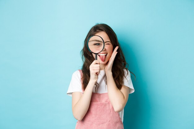 Excited young woman looks through magnifying glass, searching for someone, investigating, standing over blue background