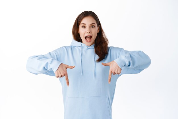 Excited young woman pointing fingers down and scream with joy, showing awesome promo deal, demonstrate advertisement on copy space, standing over white wall