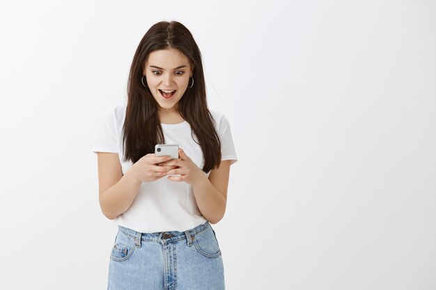 Excited young woman posing with her phone against white wall