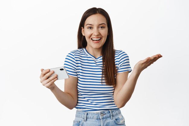 Excited young woman rejoicing, holding smartphone horizontal and spread hands sideways amazed with results, winning smth and look surprised, white background. Copy space