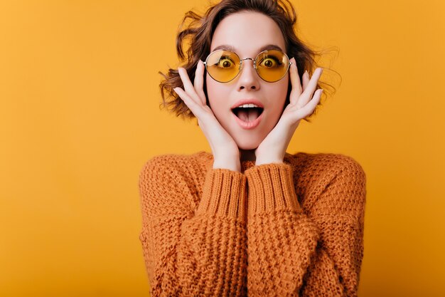 Excited young woman in round yellow glasses posing on bright space