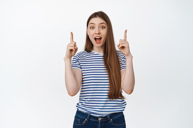 Excited young woman scream from amazement, pointing fingers up, showing sale advertisement, standing over white background