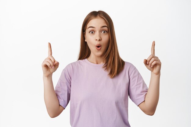 Excited young woman showing announcement, pointing fingers up and gasping amazed, impressed with discounts, standing against white wall