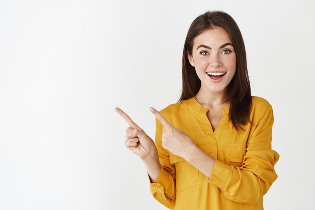 Excited young woman showing banner, pointing fingers left and smiling at camera, standing amazed at white wall