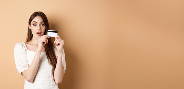 Free photo excited young woman showing plastic credit card and look amazed at camera standing on beige backgrou