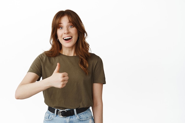 Free photo excited young woman shows thumbs up and smiles happy, give positive feedback, amazed by something cool, praise quality, say yes, like good product, standing on white