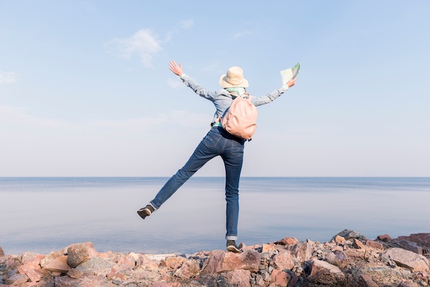 Excited young woman standing on top of rock holding map in hand overlooking the sea against blue sky