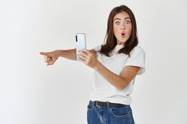 Возбужденная молодая женщина фотографирует на мобильном телефоне, указывая влево на продукт и улыбается, записывая видео на смартфон