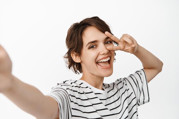 Excited young woman taking selfie with happy smile, peace v-sign on eye, standing joyful against white background