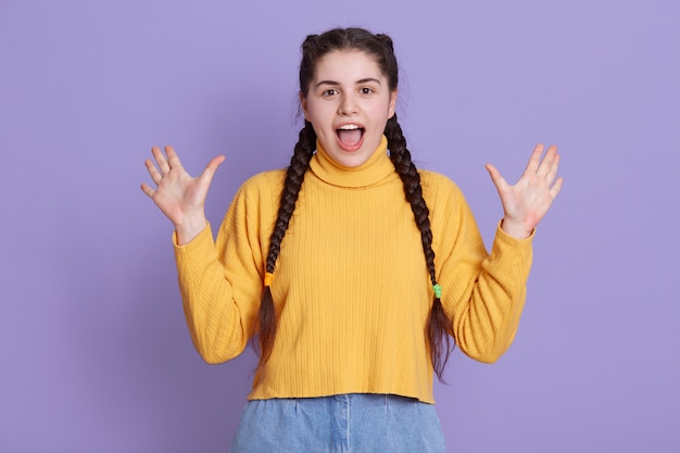 Excited young woman wearing yellow sweater, has two pigtail, posing against lilac wall