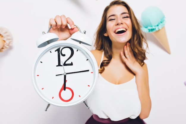 Free photo excited young woman with big clock in hand waiting for birthday party start standing on decorated wall. close-up portrait of cheerful girl rejoices at the end of the working day.