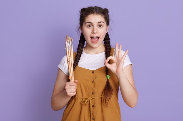 Excited young woman with painting brushed in hands, showing ok sign and yelling