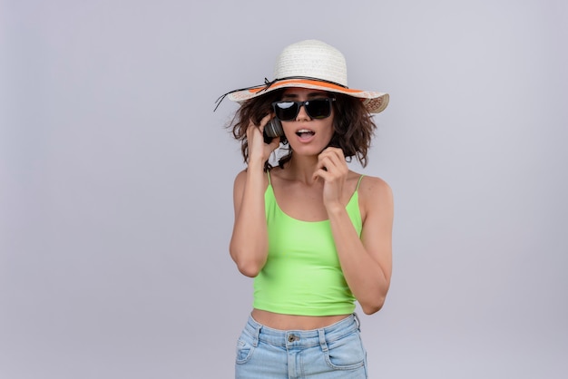 흰색 배경에 선글라스와 태양 모자를 쓰고 헤드폰을 통해 듣고 녹색 자르기 상단에 짧은 머리를 가진 흥분된 젊은 여자