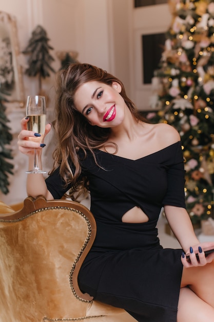 笑顔でワイングラスを育てるトレンディなメイクで興奮した若い女性。うれしそうなヨーロッパの女の子は、家でクリスマスを祝う黒いドレスを着ています。