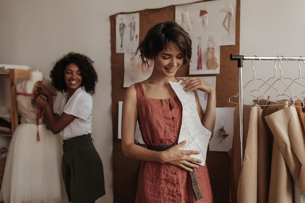 Excited young women in trendy outfits work as fashion designers Darkskinned lady designs dress Shorthaired lady looks at clothes patter
